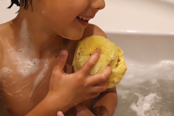 Natural sea sponge used by a child in a bathtub during bath time promotes gentle cleansing. Smiling young boy enjoying a soothing bath with frothy water and sponge.